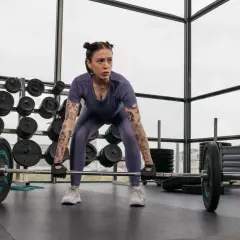 Frau mit Tattoos bereitet sich auf ein intensives Freihantel Training vor. Sie steht konzentriert vor einer Langhantel im Fitnessstudio.