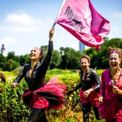 Frauen mit Schlamm und Glitzer im Gesicht rennen lachend beim Muddy Angel Run Offenbach. Eine trägt eine pinke Fahne.