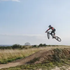 Mountainbiker springt über eine Schanze auf einer mountainbike strecke. Im Hintergrund eine Stadt und eine futuristische Skulptur.