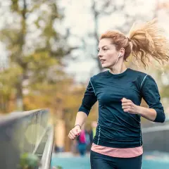 Frau mit Pferdeschwanz, die mit Kopfhörern joggen anfängt. Sie läuft auf einer Brücke, im Hintergrund Bäume und eine andere Person.