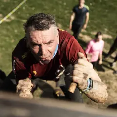 Mann kämpft sich bei einem Hindernislauf, einem echten hotfoot run, über eine Mauer. Schlamm bedeckt seine Hände und sein Trikot.