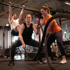Frauen und Männer trainieren im Fitnessstudio. Eine Frau macht Seilübungen, um richtig zu trainieren. Ein Mann hängt an einer Klimmzugstange.