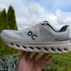 Ein weißer Cloudsurfer next Laufschuh mit "SWISS ENGINEERING" Schriftzug, gehalten in einer Hand vor grüner Natur und blauem Himmel.
