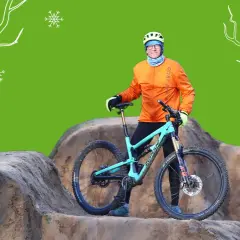 Mann mit orangefarbener Jacke und Helm steht neben seinem Mountainbike im Winter. Schneeflocken und stilisierte Bäume im Hintergrund.