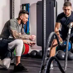 Ein Trainer kniet und erklärt einem Mann, wie er richtig trainieren im Fitnessstudio kann, während dieser mit Battle Ropes arbeitet.