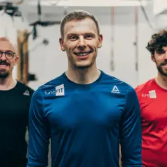Drei lächelnde Männer in Sportkleidung stehen im Fitnessstudio. Der mittlere Mann, ein fitness trainer, trägt ein blaues Shirt.