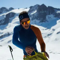 Mann in den Bergen, der sich auf das Zwiebelprinzip Kleidung vorbereitet. Er trägt eine blaue und orangefarbene Funktionsjacke und eine Skibrille.