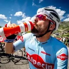 Radfahrer trinkt aus einer Flasche während eines Wettkampfs im Radsport. Er trägt ein hellblaues Trikot mit roten Logos.