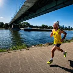Läufer mit hochrotem Kopf und erhöhtem **Puls bei Hitze** joggt unter einer Brücke am Fluss entlang.