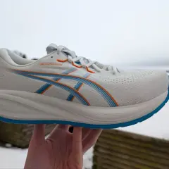 Weißer ASICS GEL-CUMULUS 26 Laufschuh mit blauen und orangen Akzenten, gehalten in einer Hand vor winterlicher Kulisse.