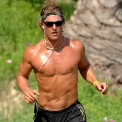 Ein Mann mit Sonnenbrille und Kopfband joggt nackt im Freien. Seine Muskeln glänzen in der Sonne, während er mit Kopfhörern Musik hört.