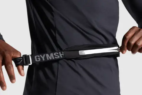 Person schließt eine Bauchtasche, ideal für das handy beim joggen. Der Gurt mit "GYMSH" Schriftzug ist verstellbar.