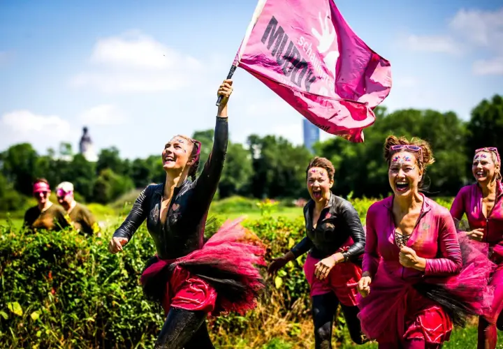 Frauen mit Schlamm und Glitzer im Gesicht rennen lachend beim Muddy Angel Run Offenbach. Eine trägt eine pinke Fahne.