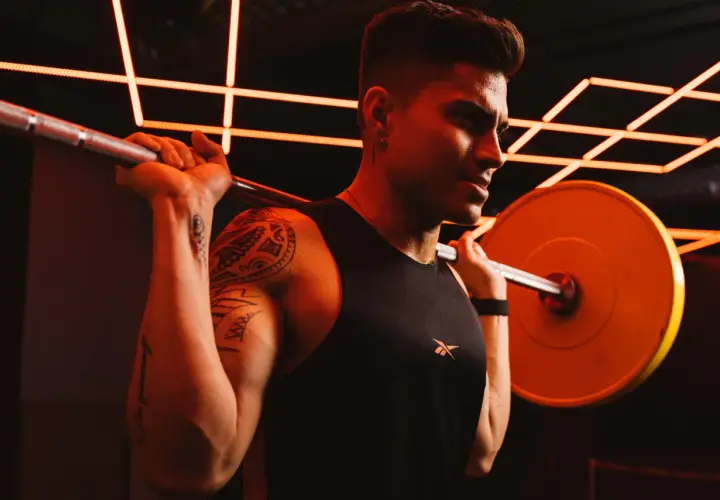 Ein Mann mit Tattoos und einem schwarzen Tanktop hebt eine Hantel. Er schwitzt und konzentriert sich auf sein Dropsatz Training.