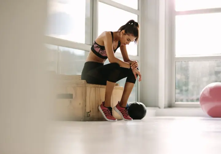 Erschöpfte Frau im **deload gym** sitzt auf einer Box, die Beine angewinkelt, der Kopf gesenkt. Ein schwarzer Medizinball und ein rosa Gymnastikball liegen daneben.