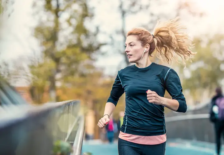 Frau mit Pferdeschwanz, die mit Kopfhörern joggen anfängt. Sie läuft auf einer Brücke, im Hintergrund Bäume und eine andere Person.