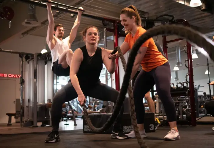 Frauen und Männer trainieren im Fitnessstudio. Eine Frau macht Seilübungen, um richtig zu trainieren. Ein Mann hängt an einer Klimmzugstange.