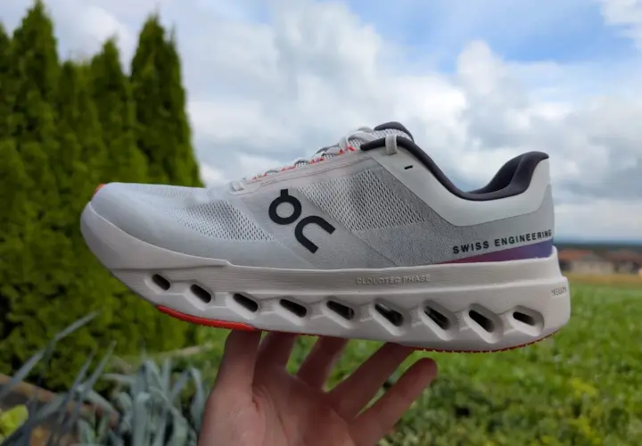 Ein weißer Cloudsurfer next Laufschuh mit "SWISS ENGINEERING" Schriftzug, gehalten in einer Hand vor grüner Natur und blauem Himmel.