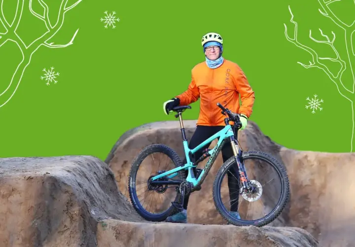 Mann mit orangefarbener Jacke und Helm steht neben seinem Mountainbike im Winter. Schneeflocken und stilisierte Bäume im Hintergrund.