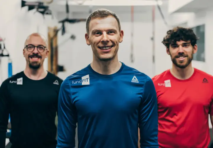 Drei lächelnde Männer in Sportkleidung stehen im Fitnessstudio. Der mittlere Mann, ein fitness trainer, trägt ein blaues Shirt.