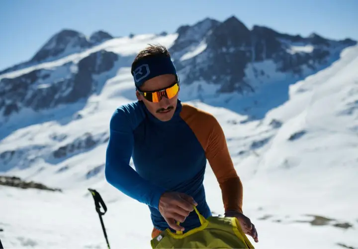 Mann in den Bergen, der sich auf das Zwiebelprinzip Kleidung vorbereitet. Er trägt eine blaue und orangefarbene Funktionsjacke und eine Skibrille.
