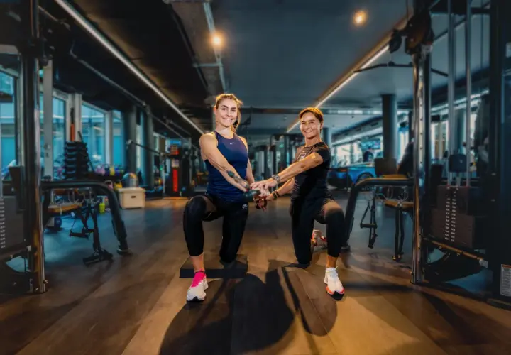 Zwei Frauen trainieren im freihantel bereich eines modernen Fitnessstudios. Sie machen eine Kniebeuge-Übung mit Kabelzug.
