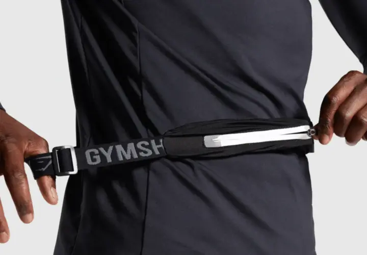 Person schließt eine Bauchtasche, ideal für das handy beim joggen. Der Gurt mit "GYMSH" Schriftzug ist verstellbar.