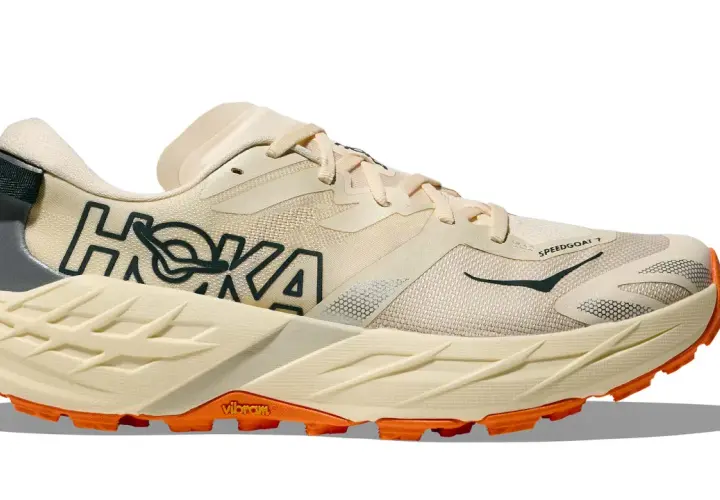 Beiger Hoka Speedgoat 7 Trailrunningschuh mit orangefarbener Vibram-Sohle und dunklem Hoka-Logo.