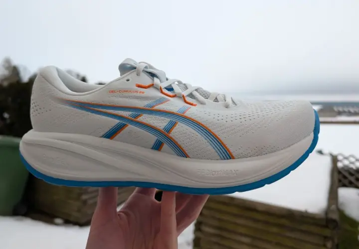 Weißer ASICS GEL-CUMULUS 26 Laufschuh mit blauen und orangen Akzenten, gehalten in einer Hand vor winterlicher Kulisse.