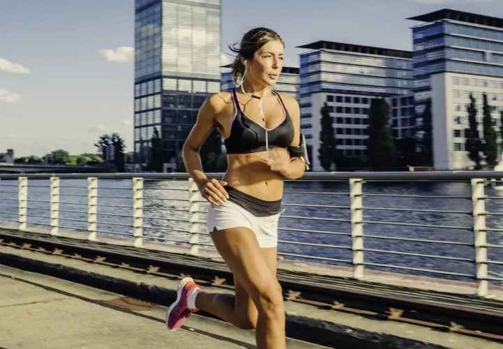 Frau läuft am Fluss entlang, umgeben von modernen Gebäuden. Sie fragt sich, wann am besten trainieren, um fit zu bleiben.
