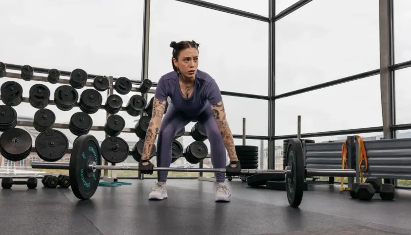 Frau mit Tattoos bereitet sich auf ein intensives Freihantel Training vor. Sie steht konzentriert vor einer Langhantel im Fitnessstudio.