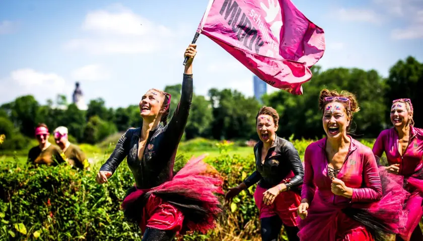 Frauen mit Schlamm und Glitzer im Gesicht rennen lachend beim Muddy Angel Run Offenbach. Eine trägt eine pinke Fahne.