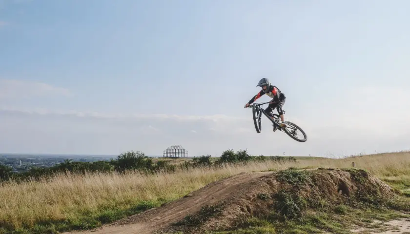 Mountainbiker springt über eine Schanze auf einer mountainbike strecke. Im Hintergrund eine Stadt und eine futuristische Skulptur.