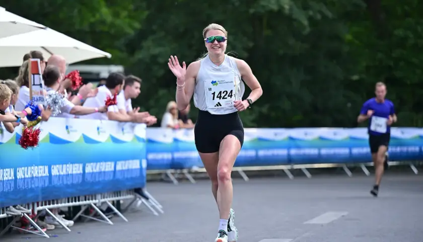 Läuferin mit Startnummer 1424 winkt fröhlich beim Berliner Teamstaffel-Lauf. Zuschauer feuern sie an.