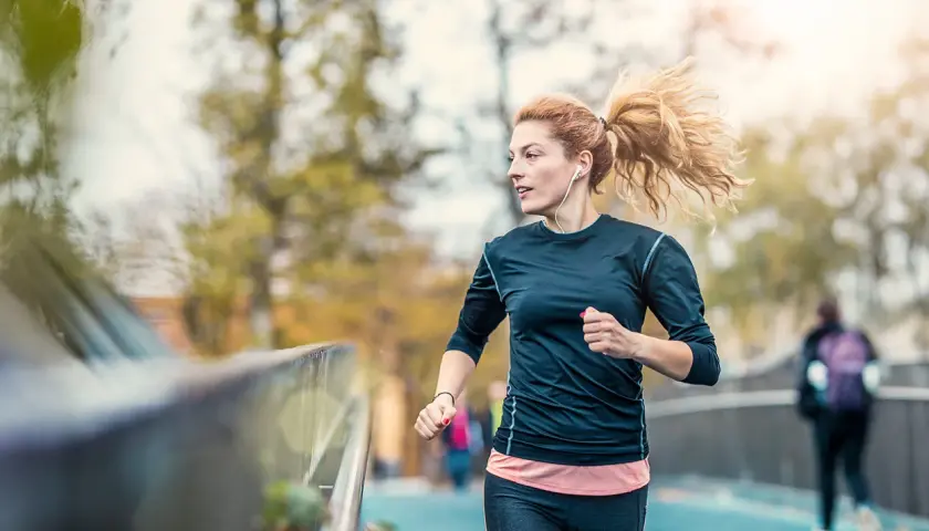 Frau mit Pferdeschwanz, die mit Kopfhörern joggen anfängt. Sie läuft auf einer Brücke, im Hintergrund Bäume und eine andere Person.