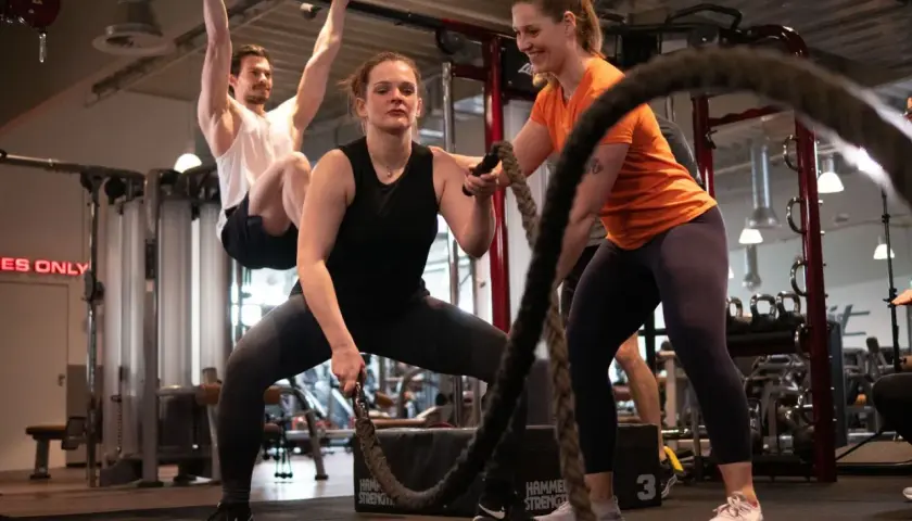 Frauen und Männer trainieren im Fitnessstudio. Eine Frau macht Seilübungen, um richtig zu trainieren. Ein Mann hängt an einer Klimmzugstange.