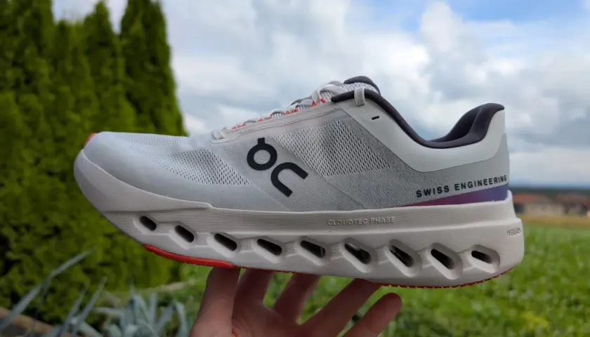 Ein weißer Cloudsurfer next Laufschuh mit "SWISS ENGINEERING" Schriftzug, gehalten in einer Hand vor grüner Natur und blauem Himmel.
