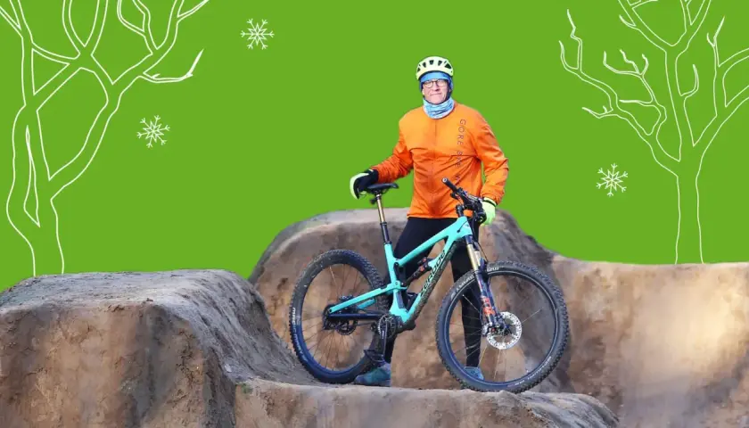 Mann mit orangefarbener Jacke und Helm steht neben seinem Mountainbike im Winter. Schneeflocken und stilisierte Bäume im Hintergrund.