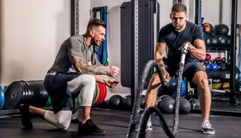 Ein Trainer kniet und erklärt einem Mann, wie er richtig trainieren im Fitnessstudio kann, während dieser mit Battle Ropes arbeitet.