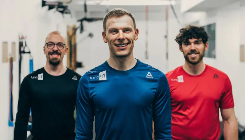 Drei lächelnde Männer in Sportkleidung stehen im Fitnessstudio. Der mittlere Mann, ein fitness trainer, trägt ein blaues Shirt.