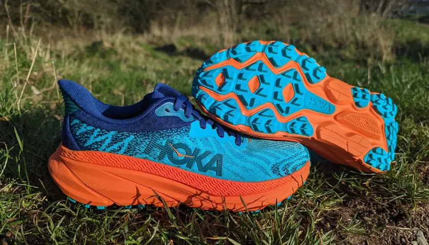 Hoka Challenger 7 Test: Blaue und orangefarbene Trailrunning-Schuhe liegen im Gras. Die Sohle zeigt ein aggressives Profil für Grip.
