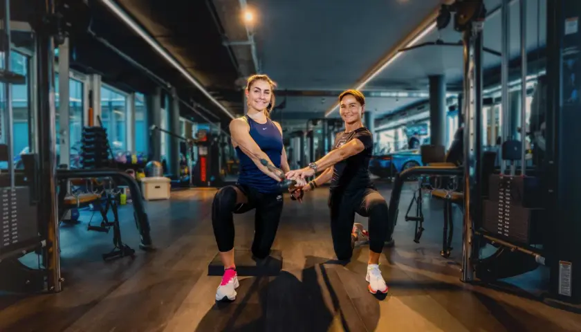 Zwei Frauen trainieren im freihantel bereich eines modernen Fitnessstudios. Sie machen eine Kniebeuge-Übung mit Kabelzug.