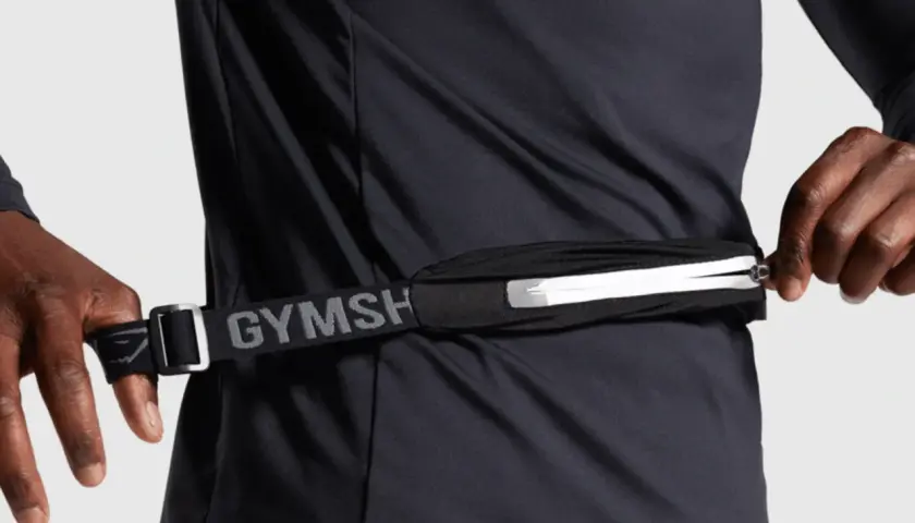 Person schließt eine Bauchtasche, ideal für das handy beim joggen. Der Gurt mit "GYMSH" Schriftzug ist verstellbar.