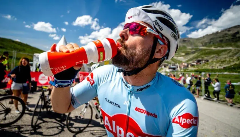 Radfahrer trinkt aus einer Flasche während eines Wettkampfs im Radsport. Er trägt ein hellblaues Trikot mit roten Logos.