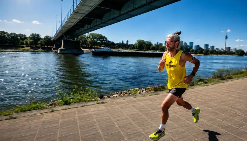 Läufer mit hochrotem Kopf und erhöhtem **Puls bei Hitze** joggt unter einer Brücke am Fluss entlang.