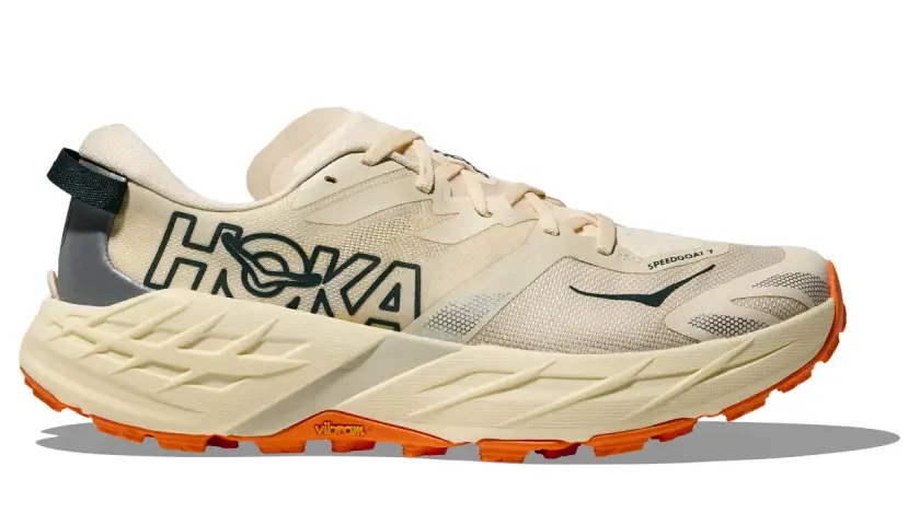 Beiger Hoka Speedgoat 7 Trailrunningschuh mit orangefarbener Vibram-Sohle und dunklem Hoka-Logo.