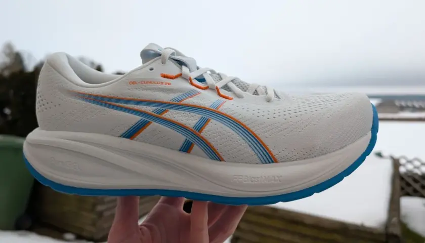 Weißer ASICS GEL-CUMULUS 26 Laufschuh mit blauen und orangen Akzenten, gehalten in einer Hand vor winterlicher Kulisse.