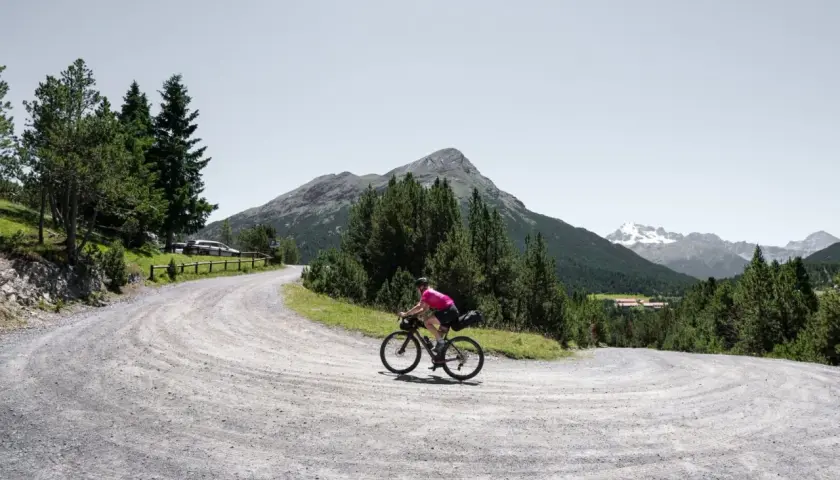 Radfahrer auf einer Schottertour in den Bergen, umgeben von Nadelbäumen und mit einem Gipfel im Hintergrund.