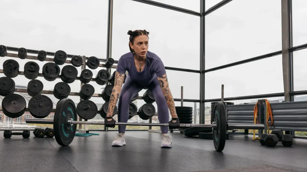 Frau mit Tattoos bereitet sich auf ein intensives Freihantel Training vor. Sie steht konzentriert vor einer Langhantel im Fitnessstudio.