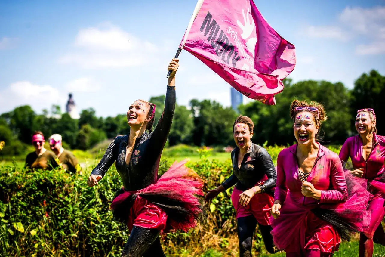 Frauen mit Schlamm und Glitzer im Gesicht rennen lachend beim Muddy Angel Run Offenbach. Eine trägt eine pinke Fahne.